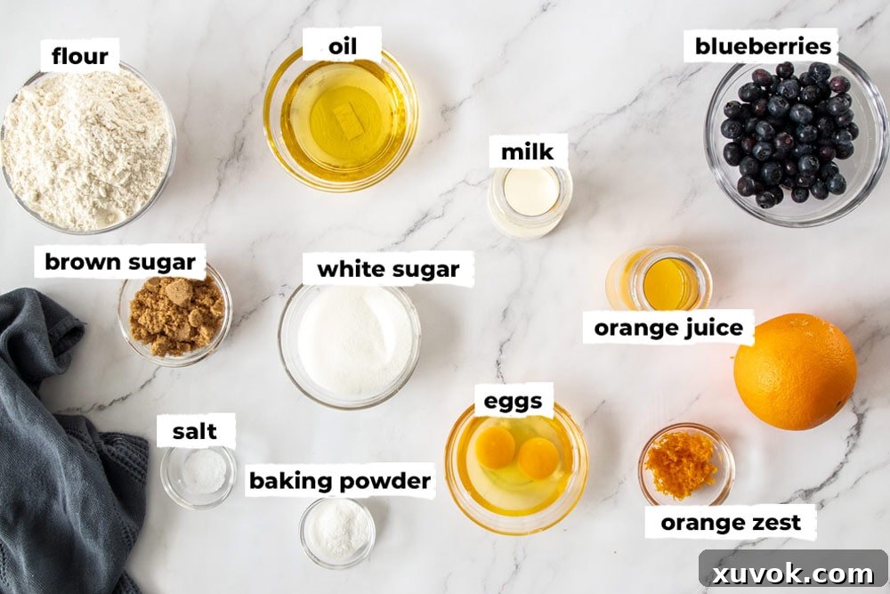 A selection of fresh ingredients laid out in glass bowls for making blueberry orange muffins, including flour, sugar, eggs, milk, blueberries, and a whole orange