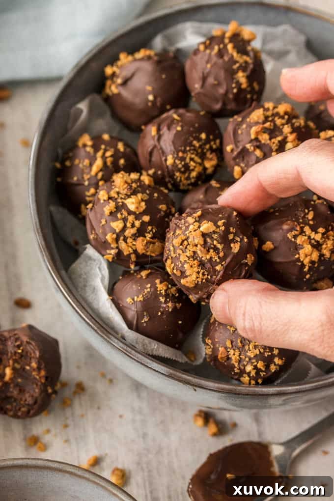 A hand picking up a chocolate truffle from a bowl, showcasing the rich, dark exterior of a homemade hazelnut praline chocolate truffle.