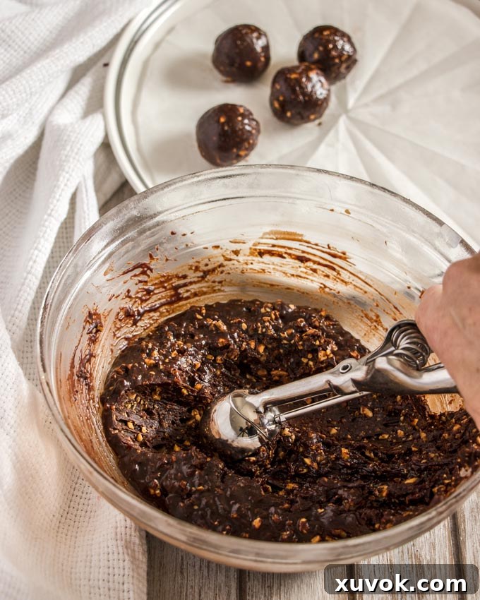 A person using a small cookie scoop to portion out hazelnut praline ganache from a bowl, preparing to roll truffles.