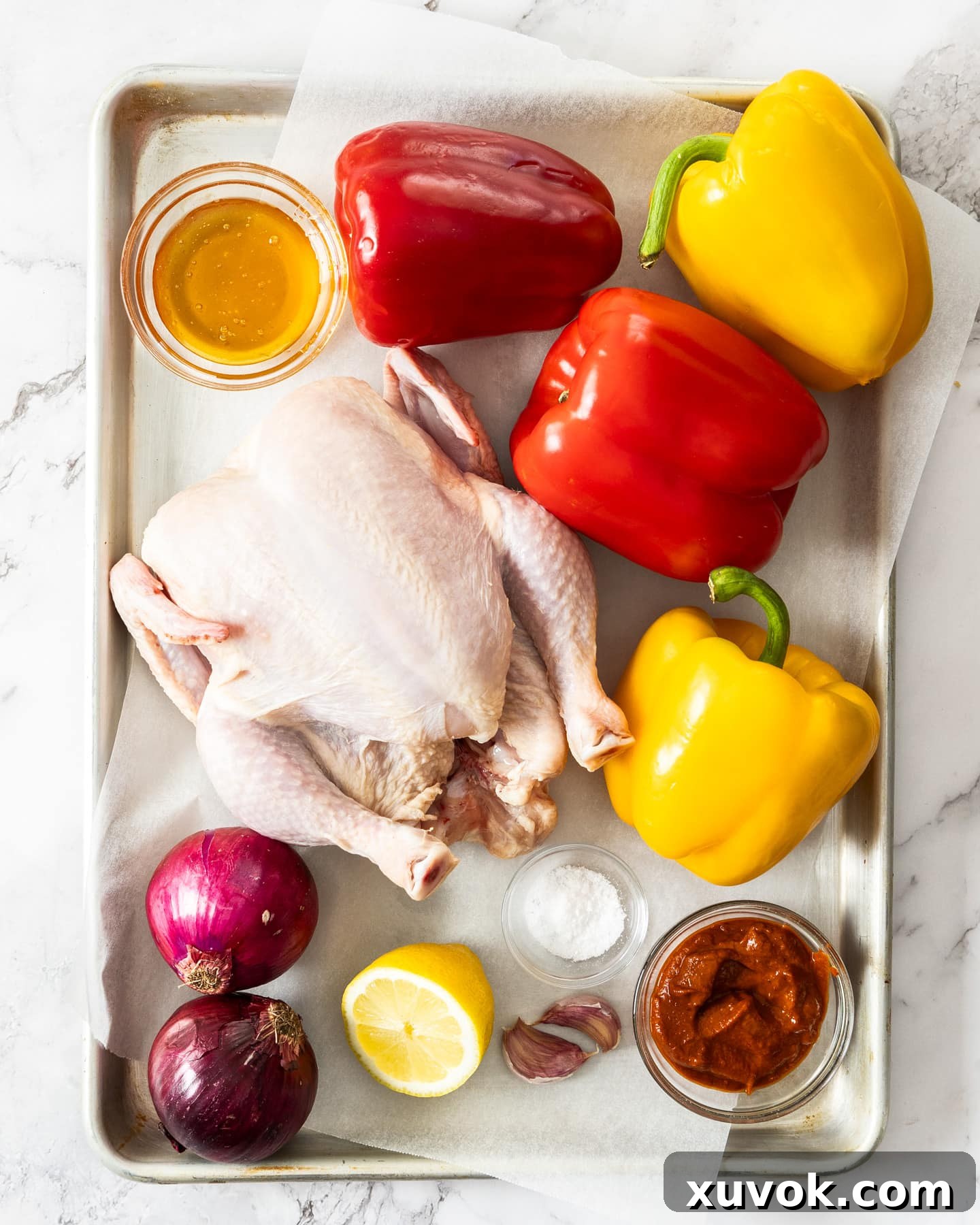 Various fresh ingredients for harissa chicken laid out on a baking tray, including a whole chicken, red onions, bell peppers, lemon, garlic, and a jar of harissa paste.