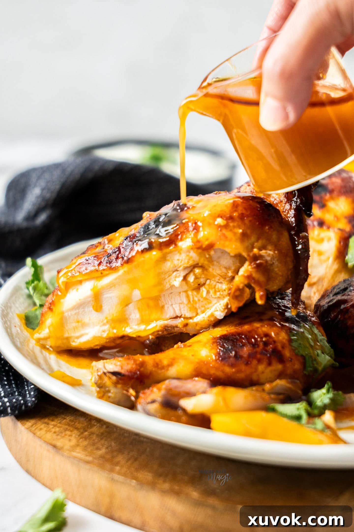 A hand pouring pan juices from a small jug over a fully roasted harissa chicken in a serving dish.