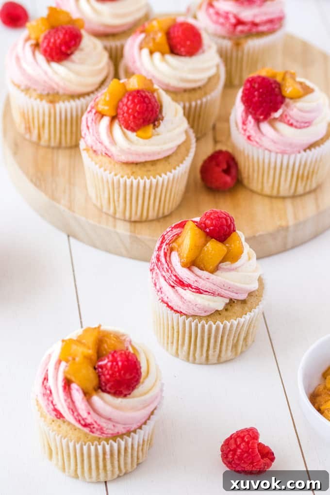 6 Peach Melba Cupcakes sitting on a round wooden board, 2 sitting on the white table top, showcasing their beautiful frosting.