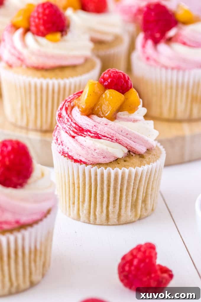 Closeup shot of a Peach Melba Cupcake, with a vibrant three-tone frosting and a peach slice on top, highlighting its beauty.