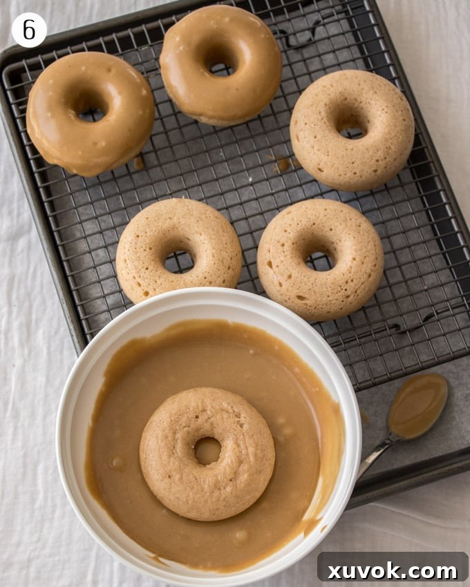A photo showing one cake donut being dipped in the brown butter glaze, with others on a cooling rack.
