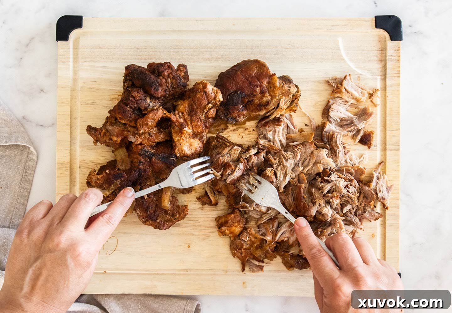 Hands holding two forks, shredding pork on a wooden chopping board.