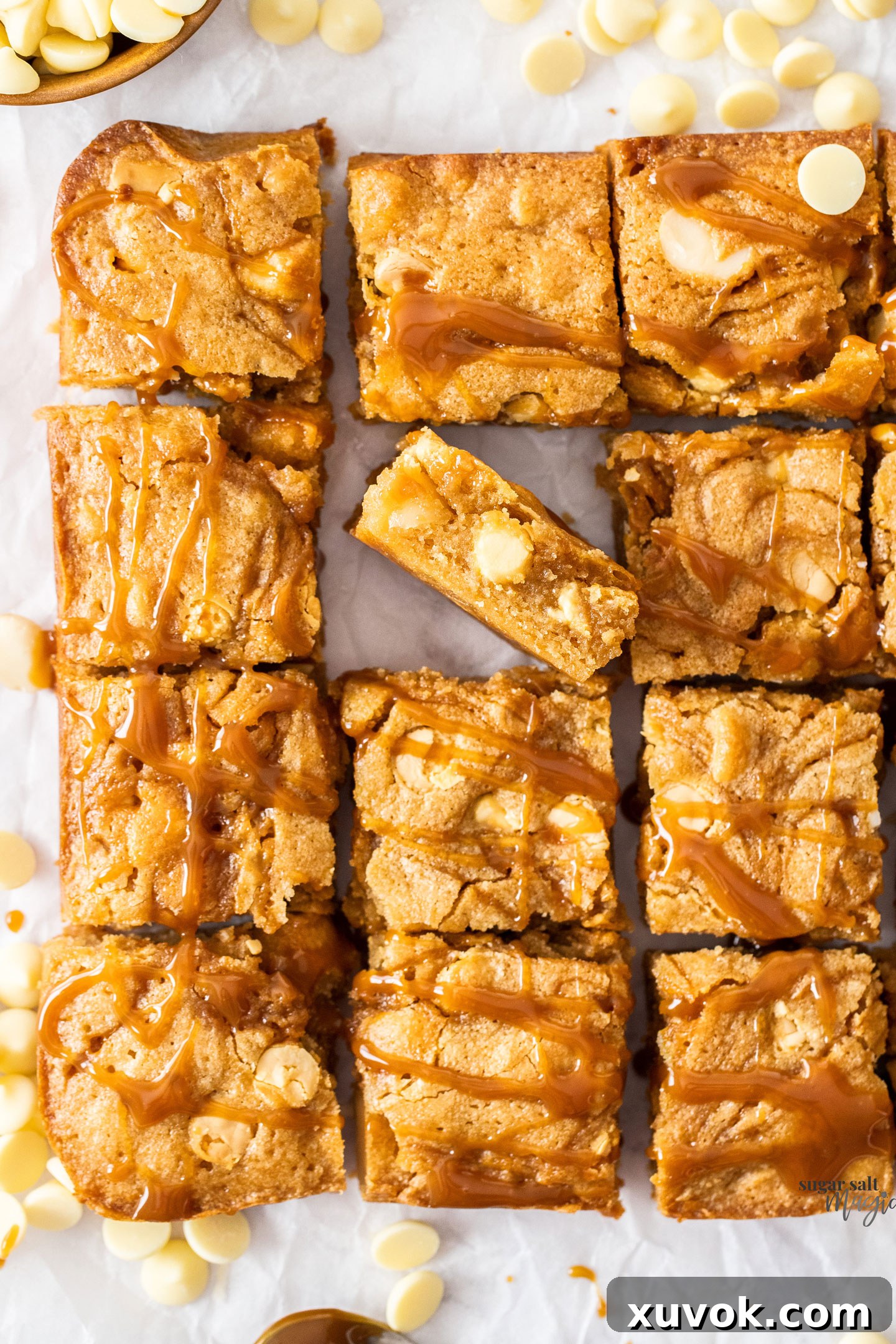 A stunning arrangement of twelve gooey caramel blondies, with one blondie standing upright to reveal its irresistible soft, fudgy, and caramel-swirled interior, adorned with white chocolate and macadamia nuts.