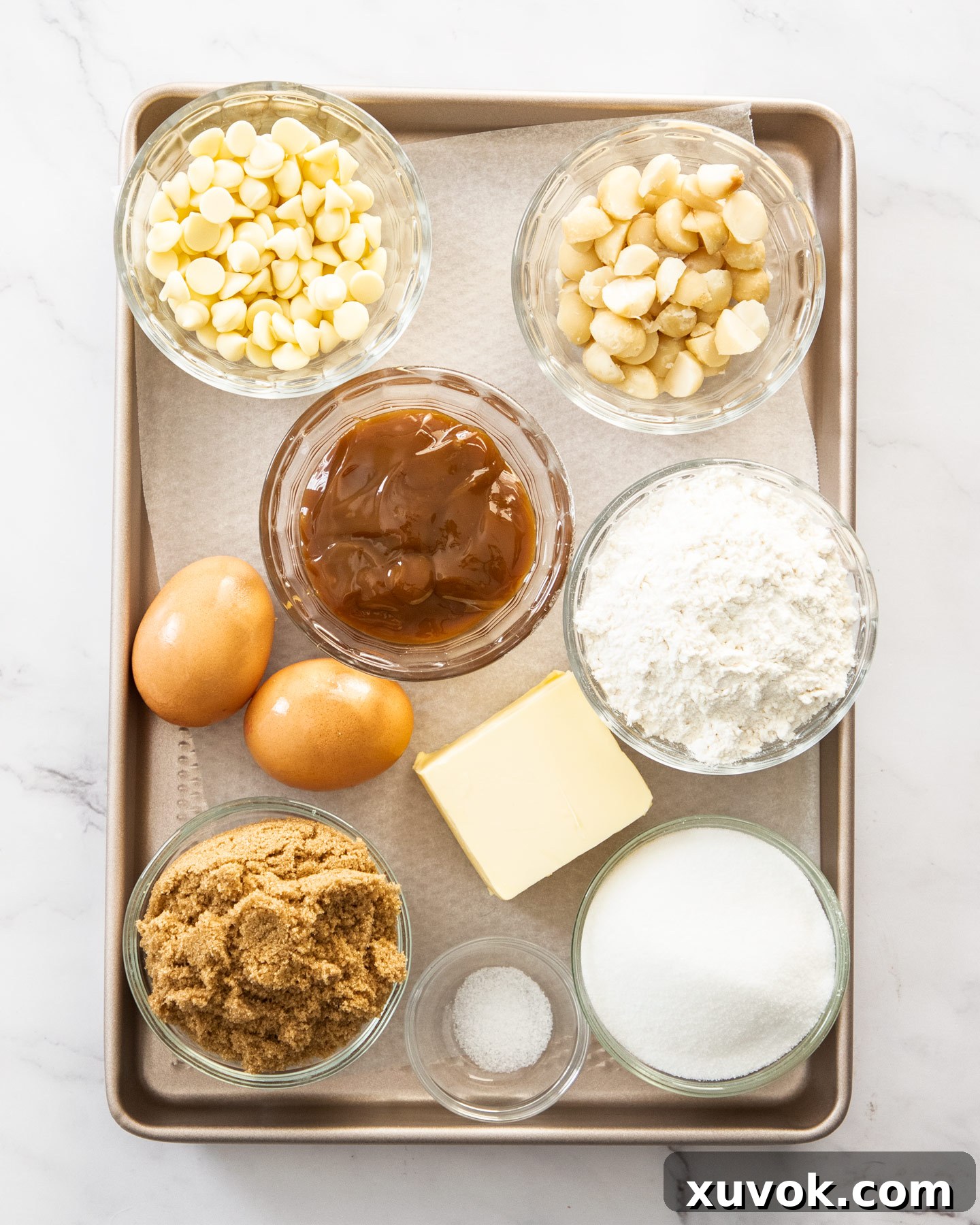 A selection of fresh ingredients for caramel blondies, neatly arranged on a rustic baking tray, featuring butter, eggs, flour, sugars, caramel sauce, white chocolate chips, and macadamia nuts.