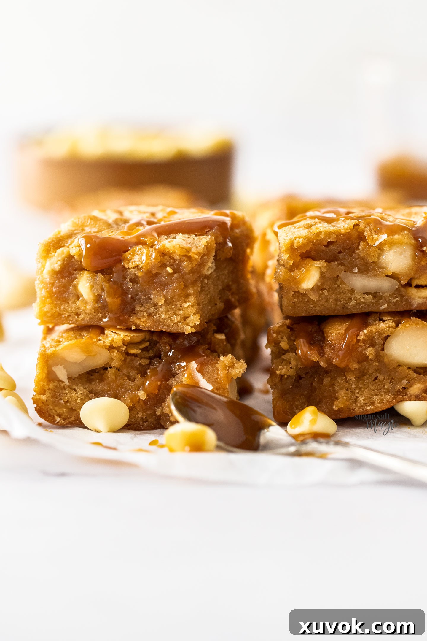 Two enticing stacks of caramel blondies, each perfectly square and rich, sitting beside a scattering of creamy white chocolate chips, highlighting their indulgent ingredients.