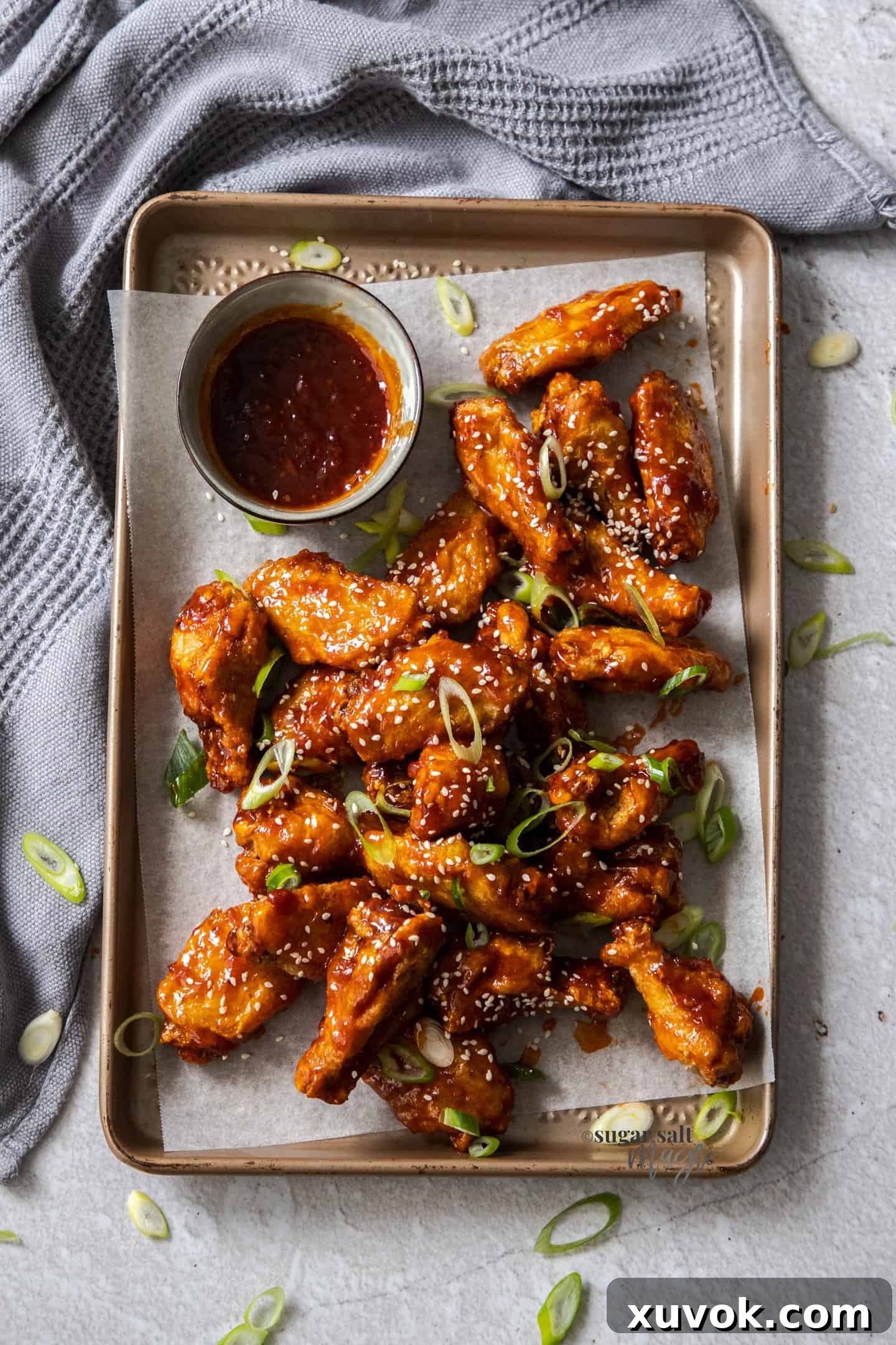 Top down view of a pile of fried sticky chicken wings on a baking tray, garnished with sesame seeds and spring onions.