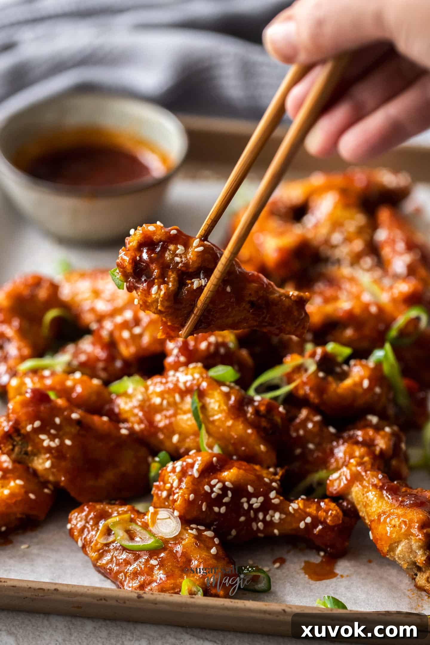 A chicken wing drumette held by chopsticks, with a pile of glazed wings in the background.