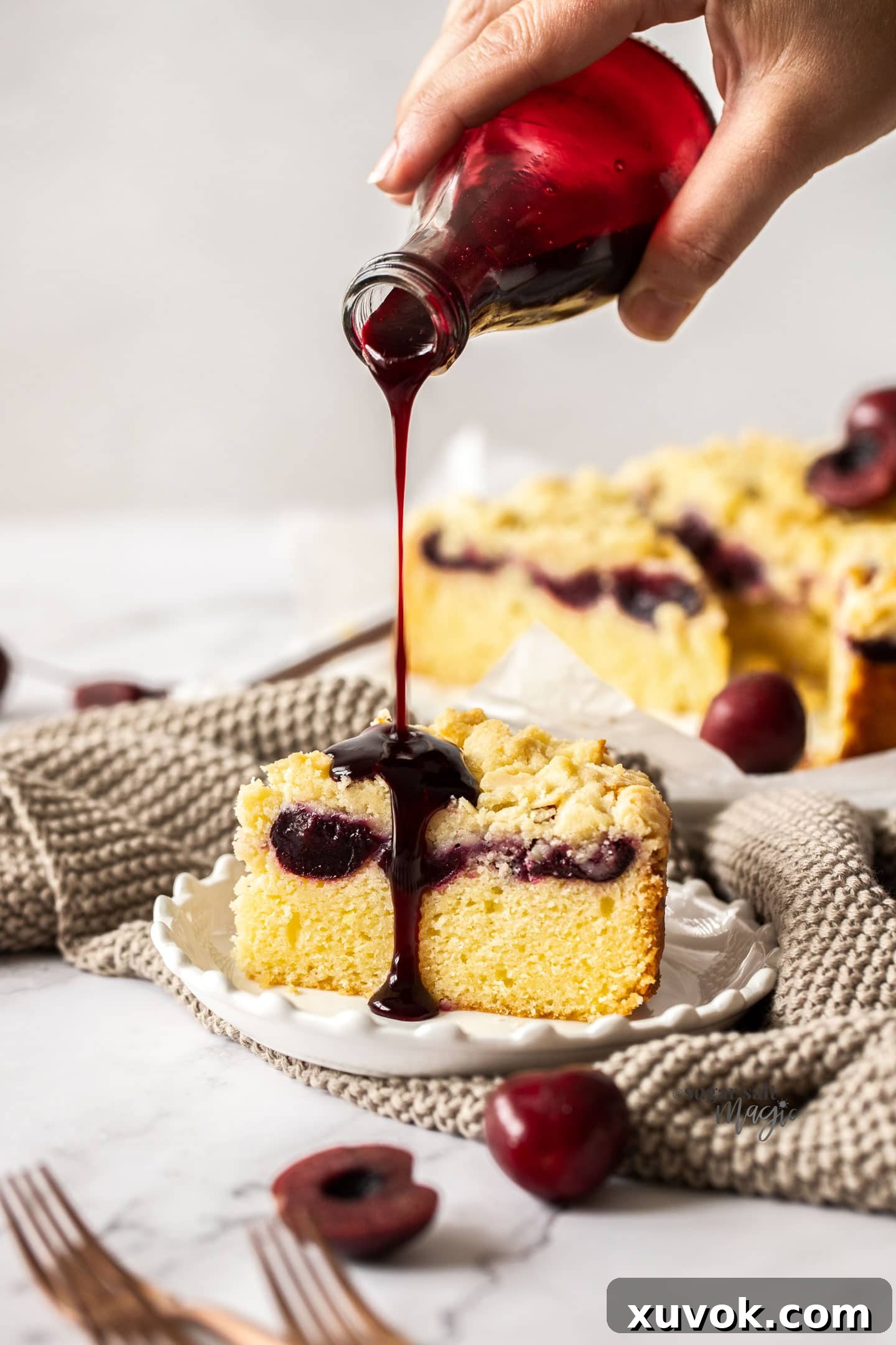 A small bottle of cherry syrup being poured artfully over a fresh slice of almond cherry crumb cake on a plate.