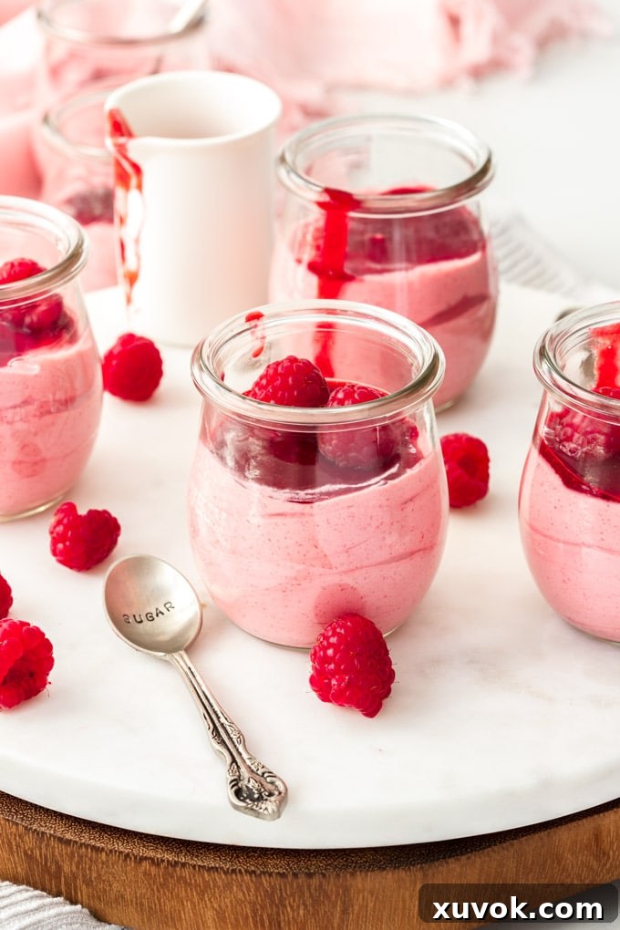A white platter holding 4 small glass jars with raspberry mousse. Fresh raspberries scattered around