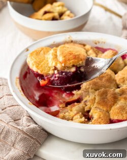 A spoon filled with warm apple and plum cobbler, scooping from a white pie dish.