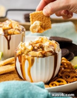 A chocolate cookie dipped into a white bowl of Snickers Dip, with other cookies and pretzels nearby, ready for a party.