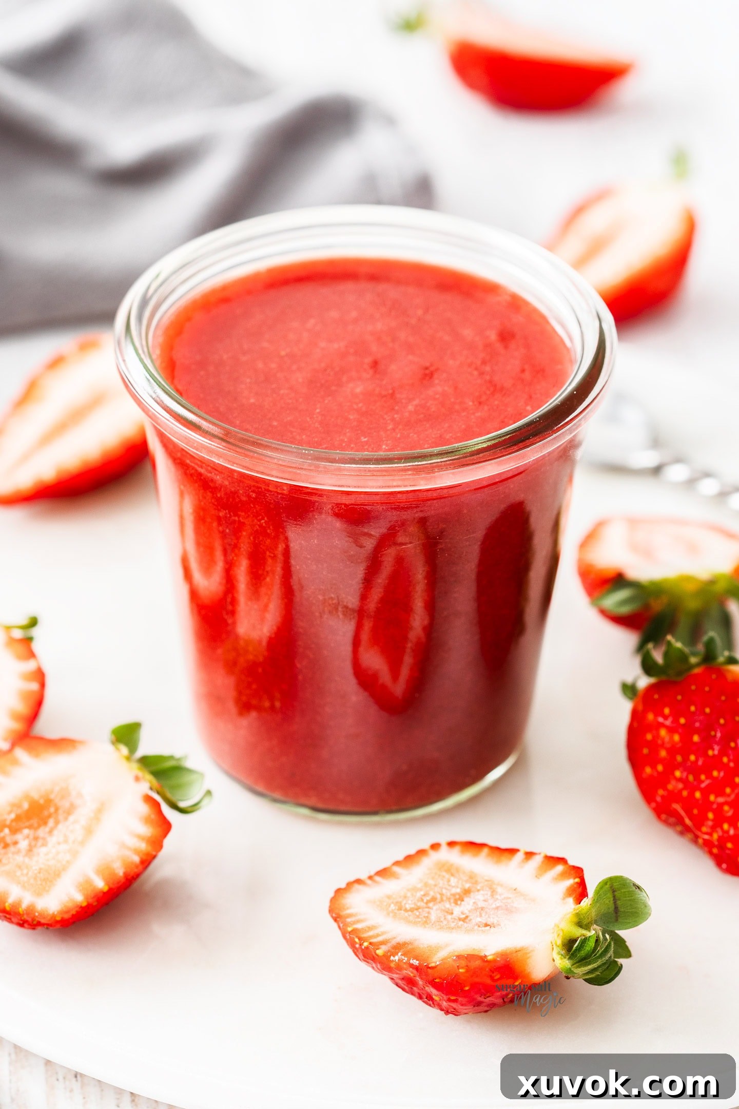 A glass pot filled with strawberry sauce surrounded by strawberries.