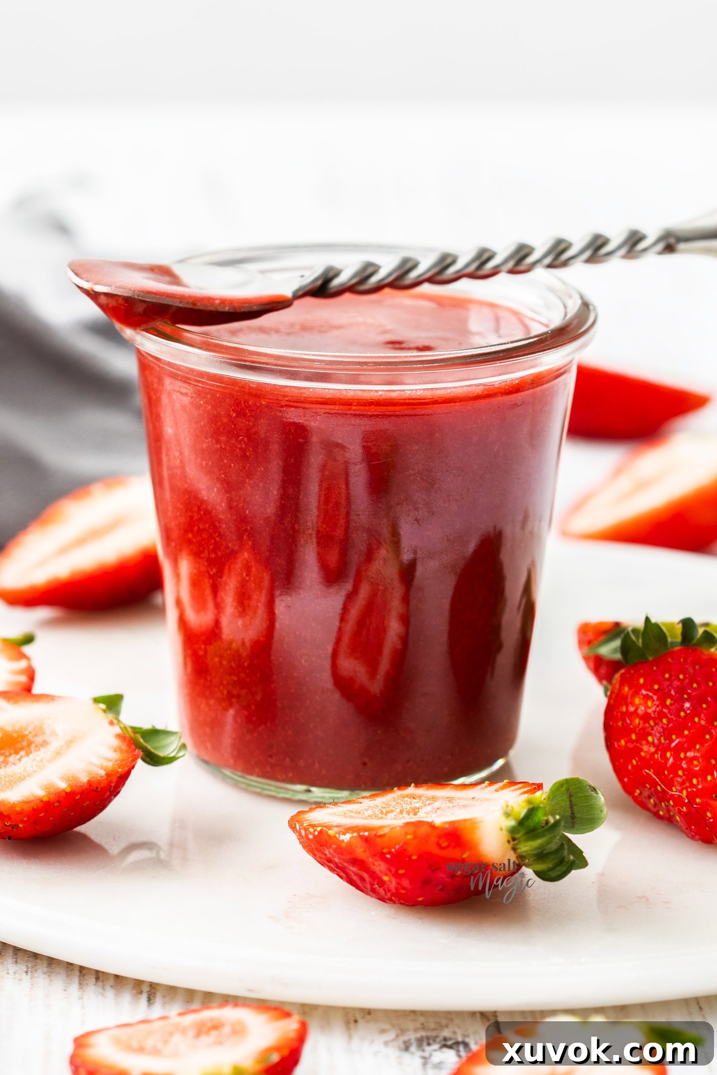 A glass pot filled with strawberry sauce with a spoon resting on top and surrounded by strawberries.