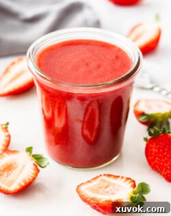 A glass pot filled with strawberry sauce surrounded by strawberries.