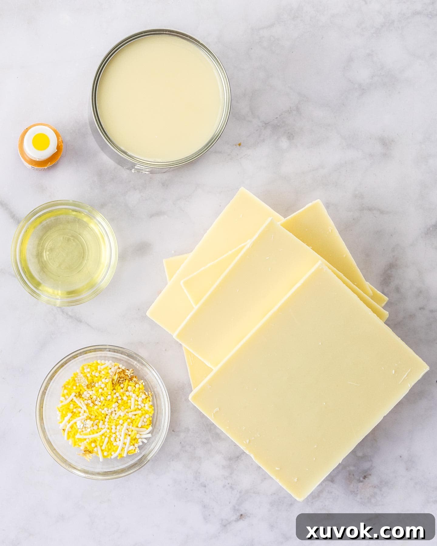 A close-up view of the ingredients for lemon fudge, including condensed milk, white chocolate, lemon extract, and yellow food coloring, laid out on a clean surface.
