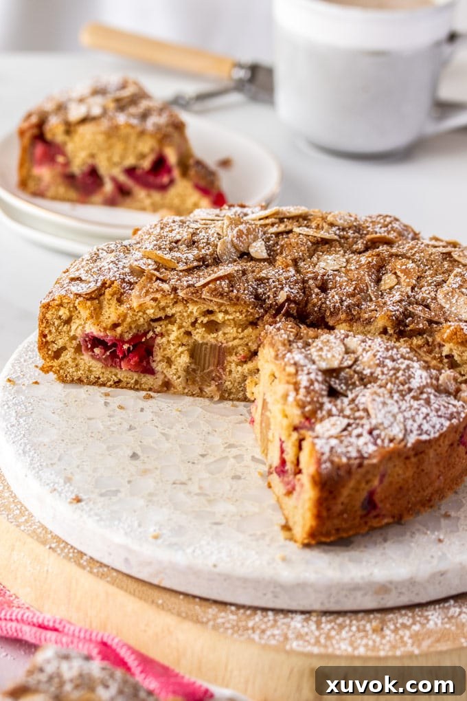 A beautifully baked sour cream rhubarb coffee cake with several slices removed, showcasing its fluffy texture and pink rhubarb chunks. It rests on an elegant marble cake platter, inviting a cozy tea-time moment.