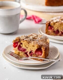 A slice of sour cream rhubarb coffee cake sitting on a white plate with a fork next to it, and a cup of coffee in the background, inviting a delicious moment.