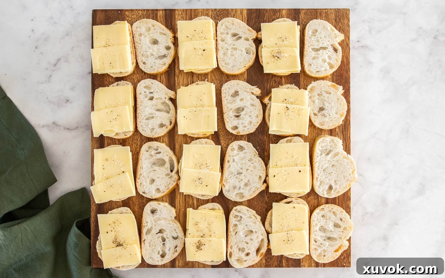Rows of thinly sliced baguette rounds, with half of them topped with layered cheddar and gouda cheese, on a rustic wooden board.