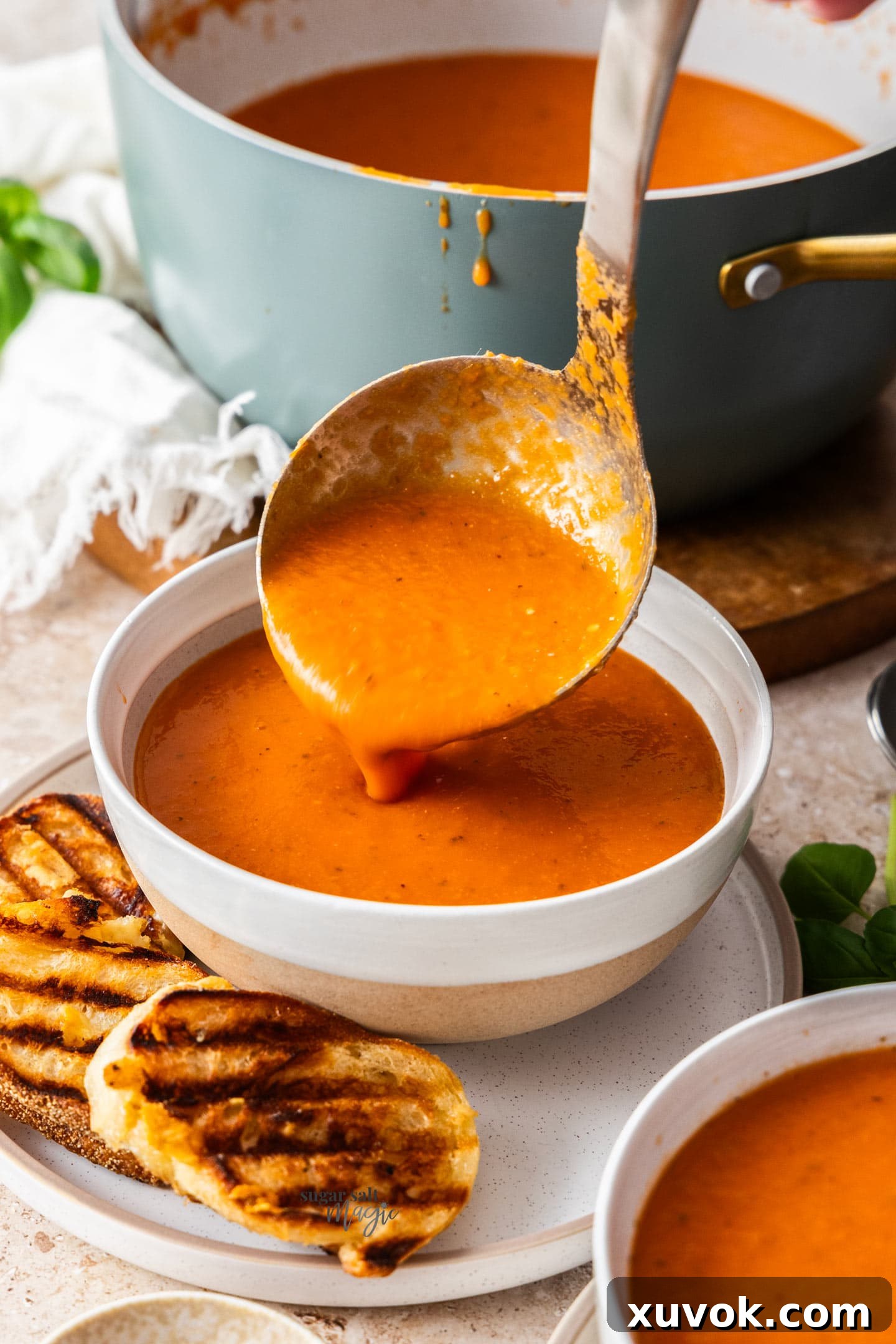 A ladle gracefully pouring rich, red roasted garlic tomato soup into a rustic white bowl, ready for serving.