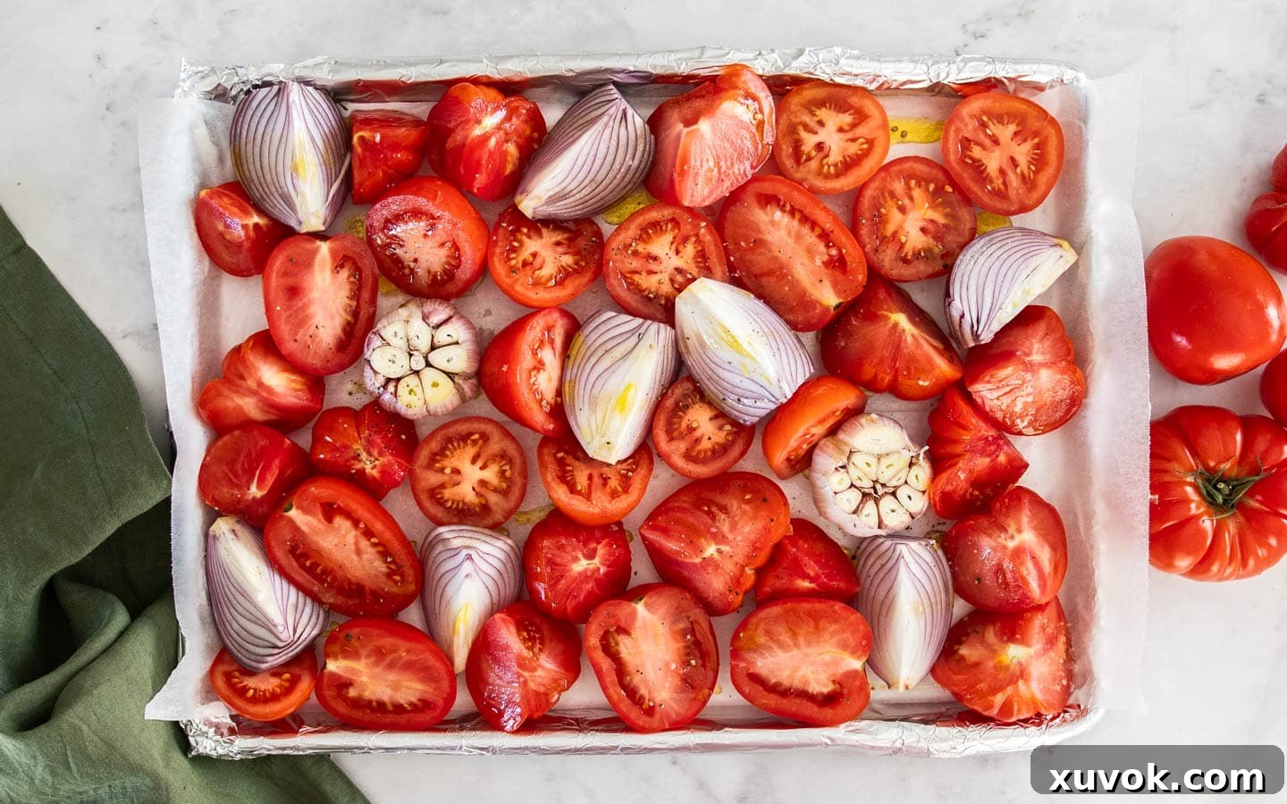A baking sheet filled with freshly chopped tomatoes, quartered red onions, and two garlic bulbs with their tops sliced off, all drizzled with olive oil and seasoned.