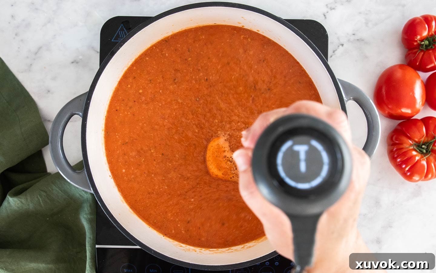 A hand holding an immersion blender, blending the roasted vegetables and stock in a Dutch oven until smooth.