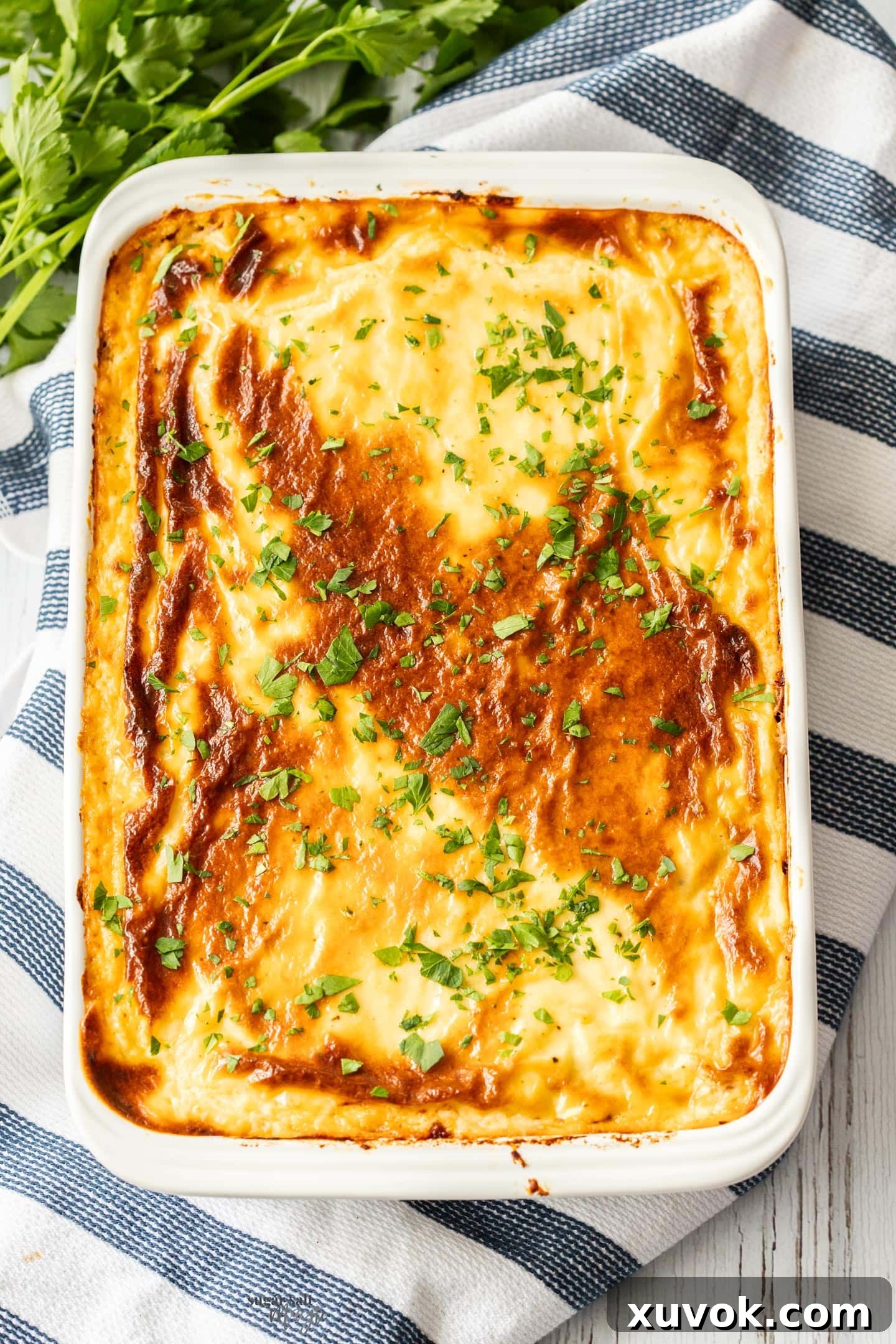 Top down view of a baked pastitsio on a striped tea towel. A golden brown bechamel topping with a crisp crust, revealing layers beneath.