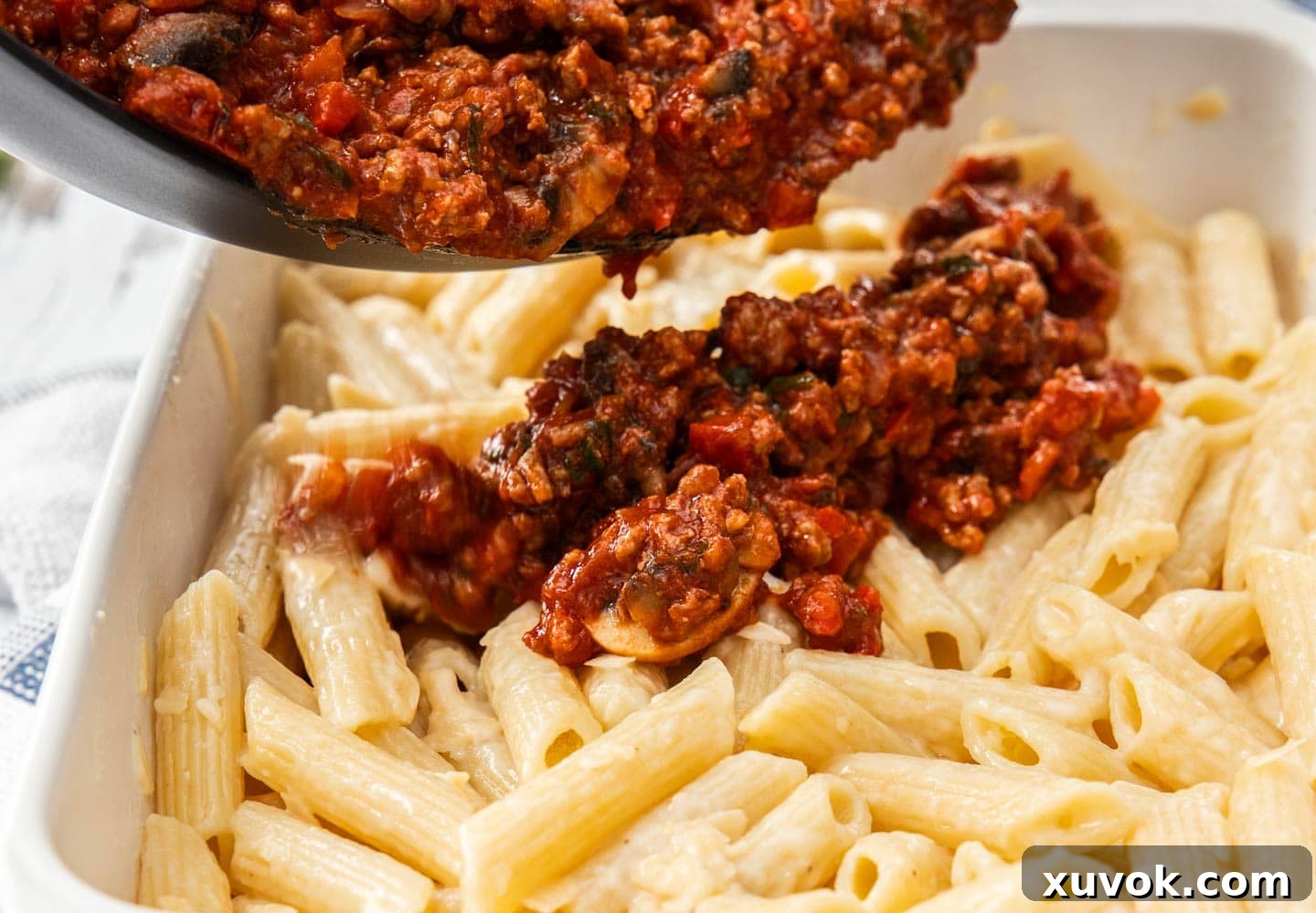 The seasoned meat sauce being poured over the prepared pasta layer in the baking dish.