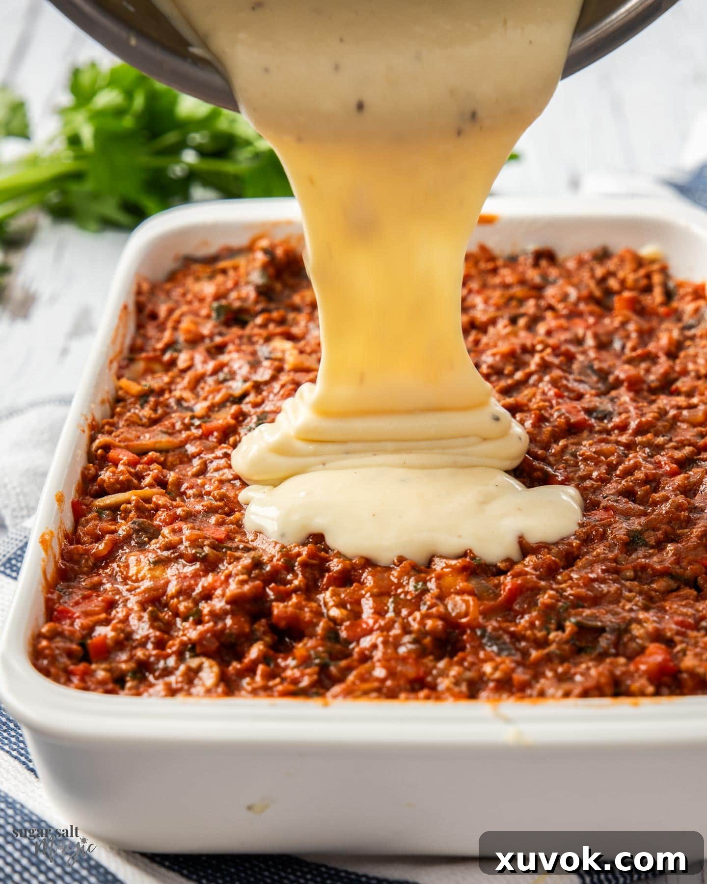 Thick béchamel sauce being poured and spread evenly over the meat filling in the Pastitsio, ready for baking.