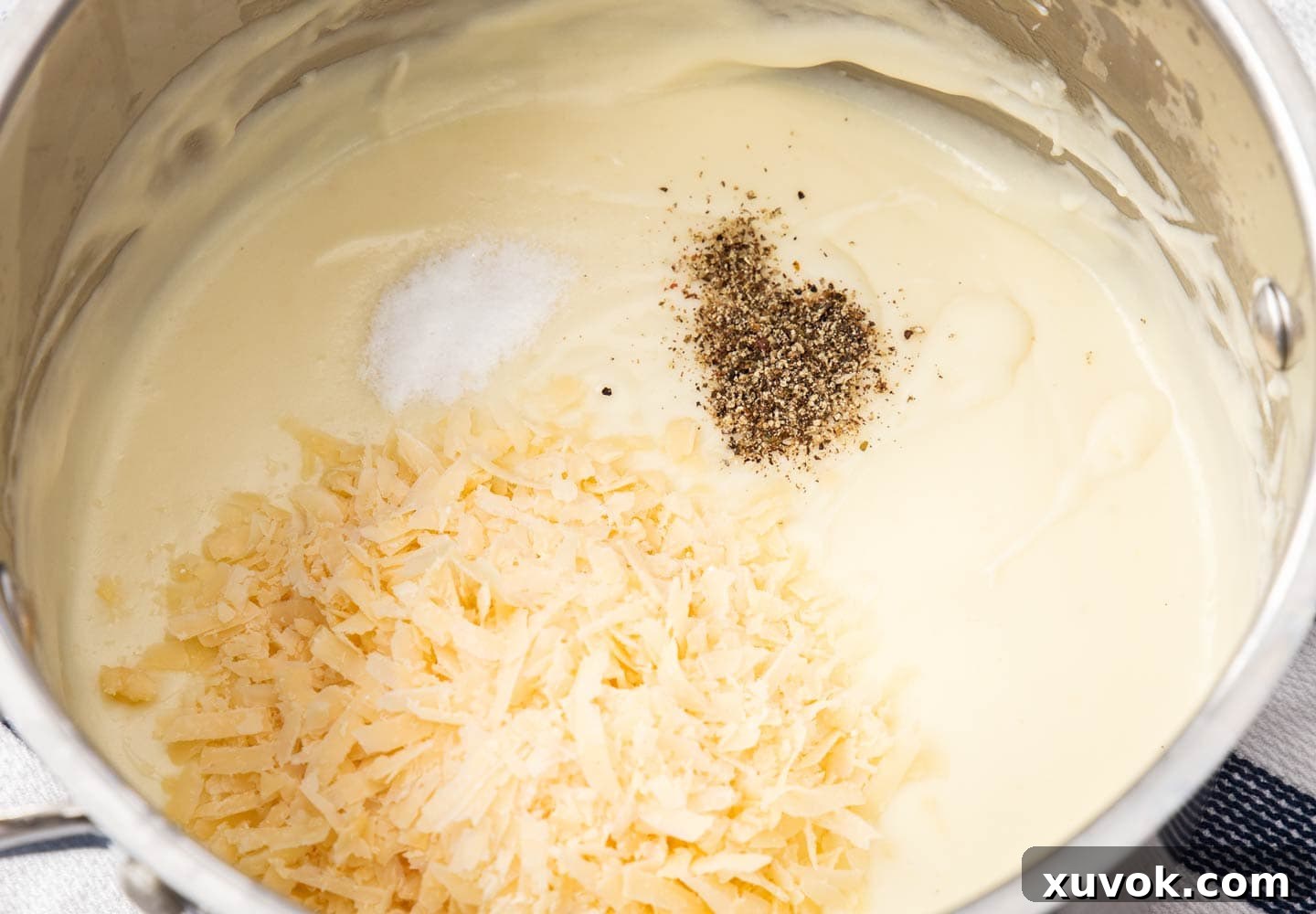 The smooth béchamel sauce in a saucepan, with salt, pepper, and grated cheese sprinkled on top, awaiting a final stir.