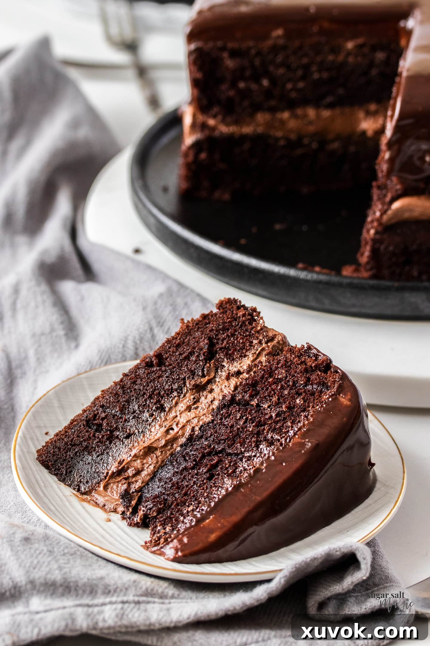 A tempting slice of Black Magic Cake, showcasing its deep, rich chocolate color, moist texture, and layers of luscious ganache buttercream, rests on a pristine white dessert plate. The perfect treat ready to be enjoyed.