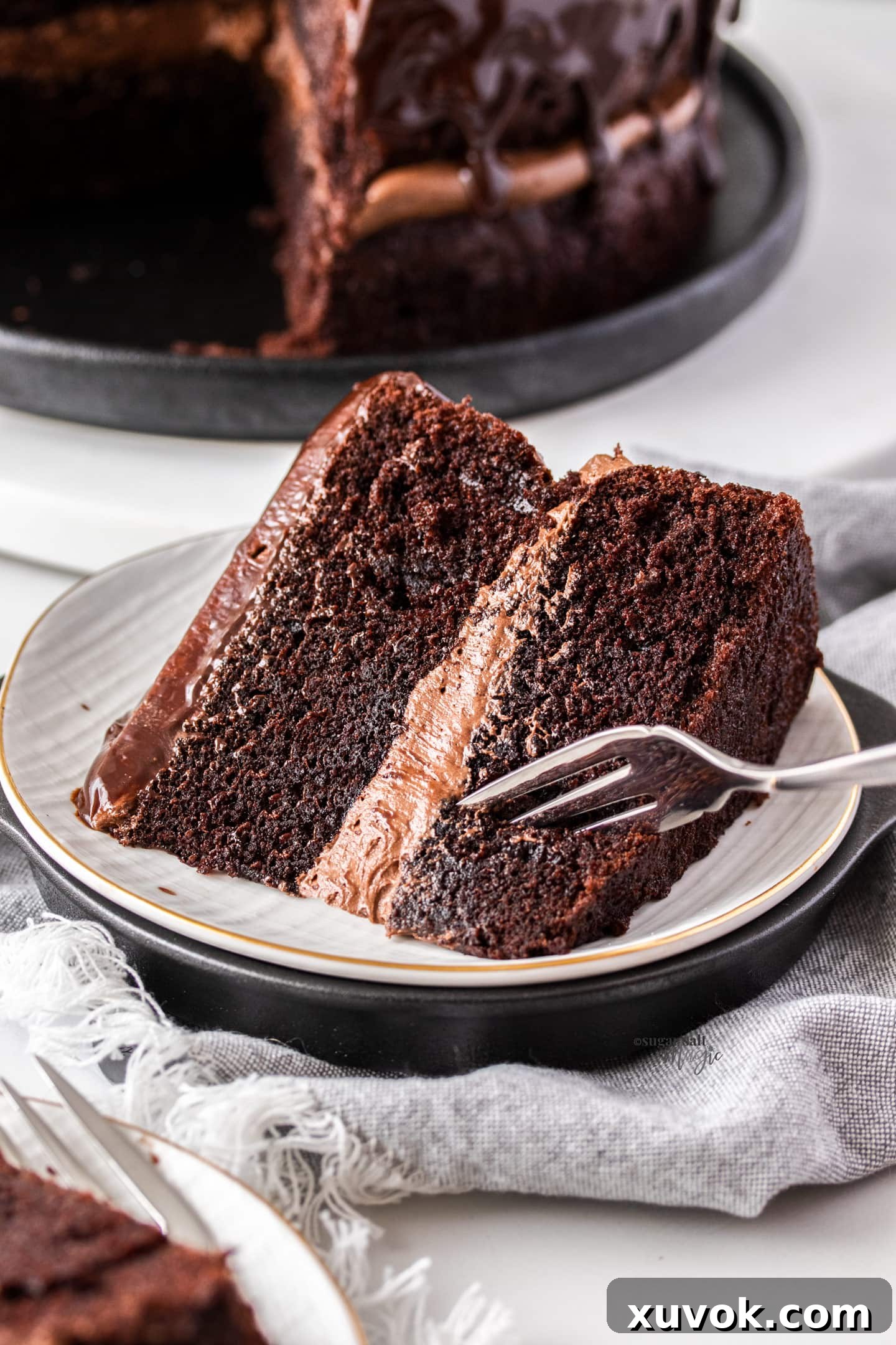 A fork gently digs into a generous slice of Black Magic Cake, revealing its moist, dark crumb and layers of glossy chocolate ganache. The image captures the irresistible texture and richness of the cake.