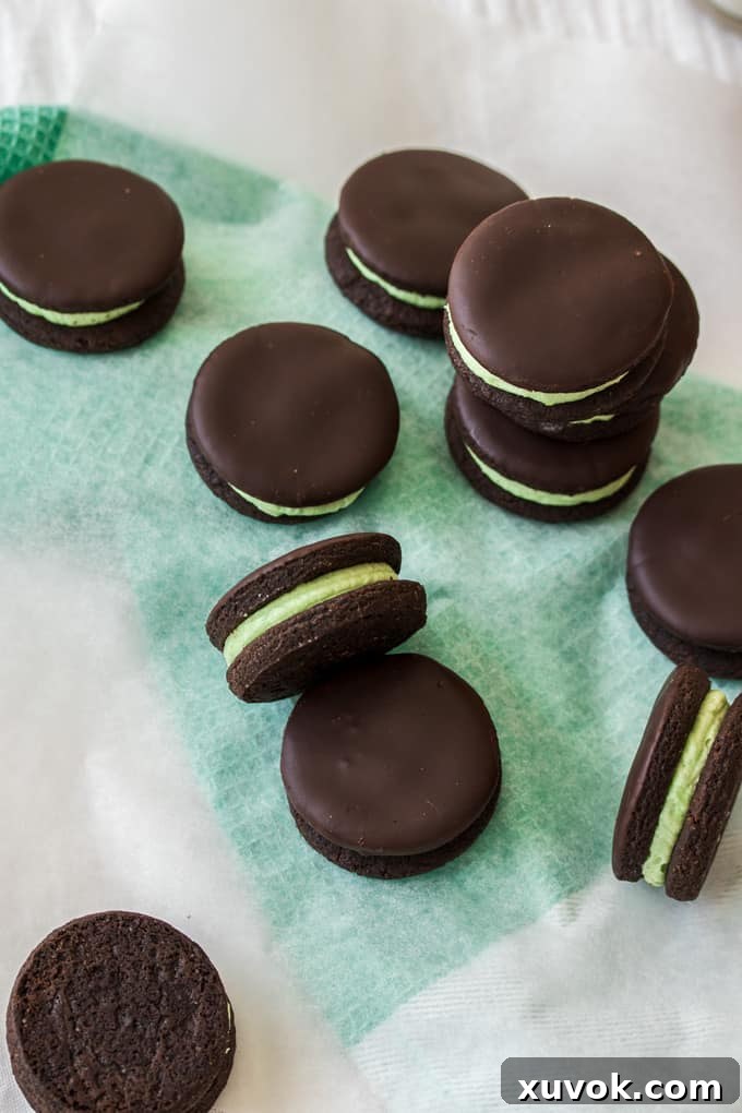 Crisp dark chocolate cookies with a vibrant green peppermint buttercream filling, elegantly arranged on a sheet of white baking paper over a green towel. Perfect for a festive snack or dessert.