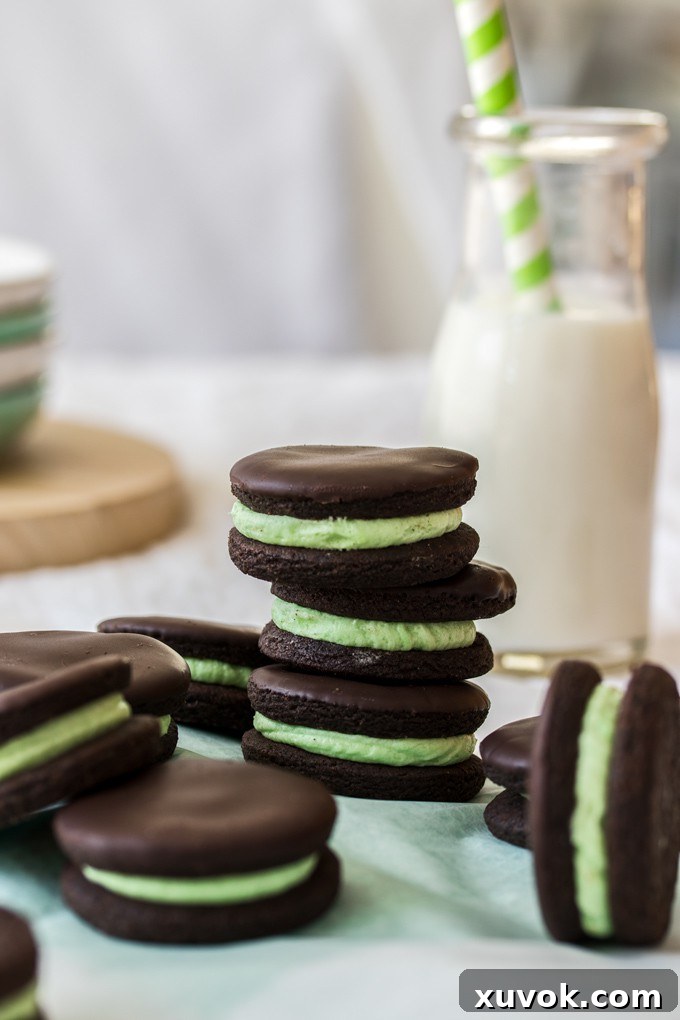 A charming stack of three dark chocolate cookies, each generously filled with a vibrant green peppermint buttercream, stands beside a rustic bottle of fresh milk. This inviting image suggests a perfect afternoon treat.