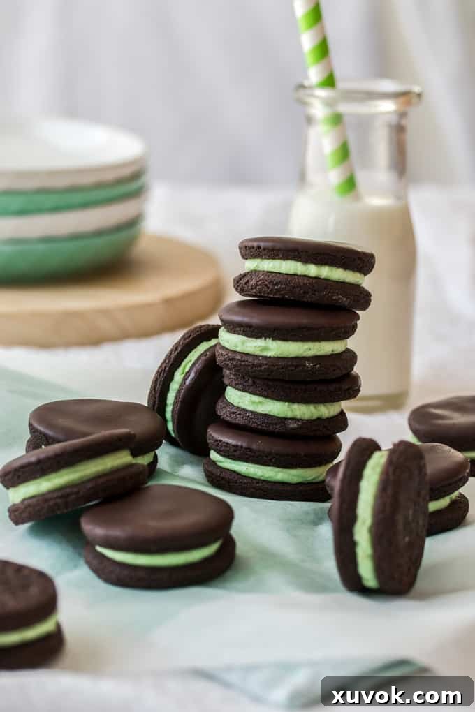 A captivating stack of four dark chocolate cookies, each generously filled with bright green peppermint buttercream, with a bottle of fresh milk subtly blurred in the background. This image highlights the irresistible appeal of these homemade treats.