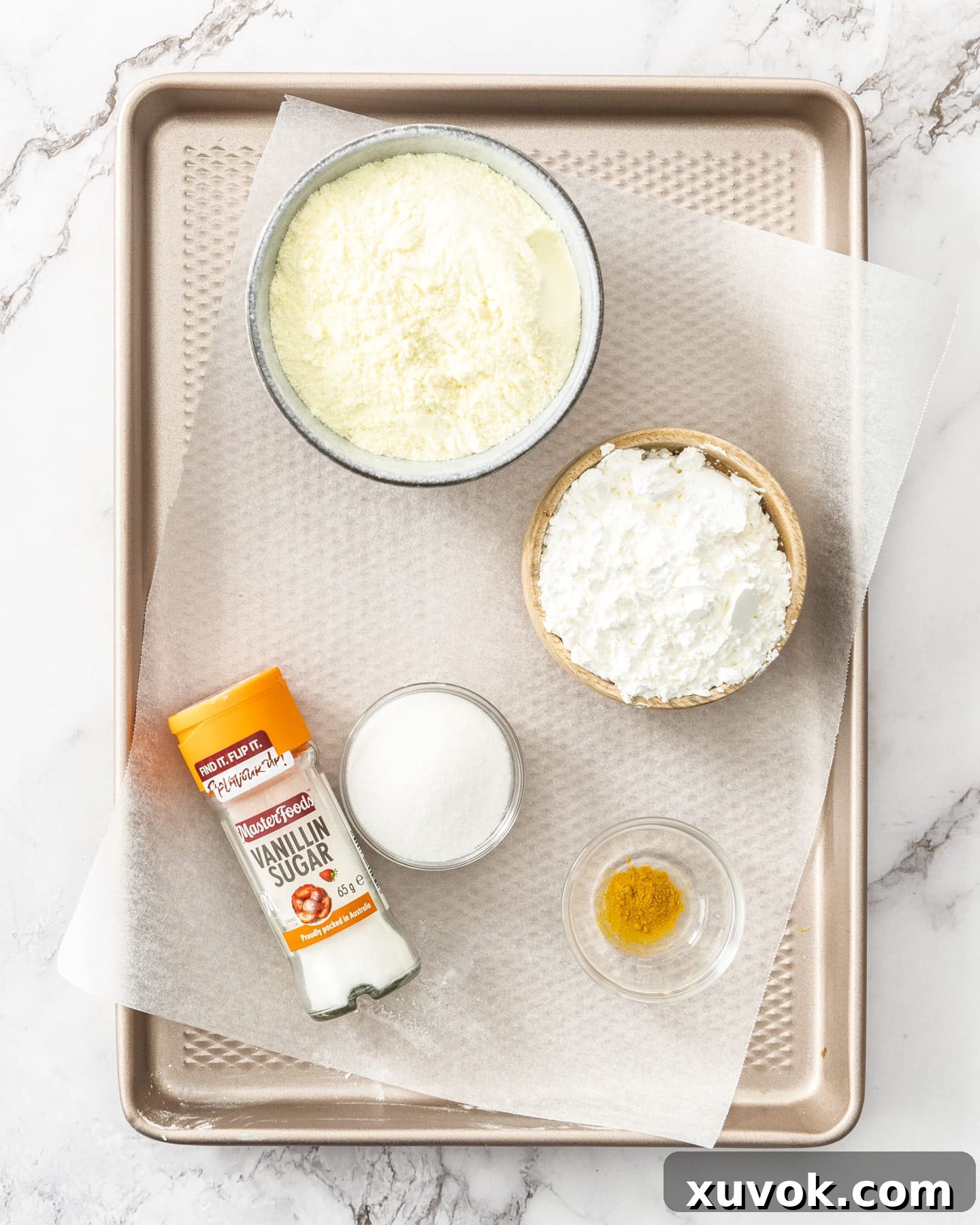 Individual ingredients for custard powder, including milk powder, cornflour, vanilla sugar, and yellow food coloring, laid out on a baking tray.