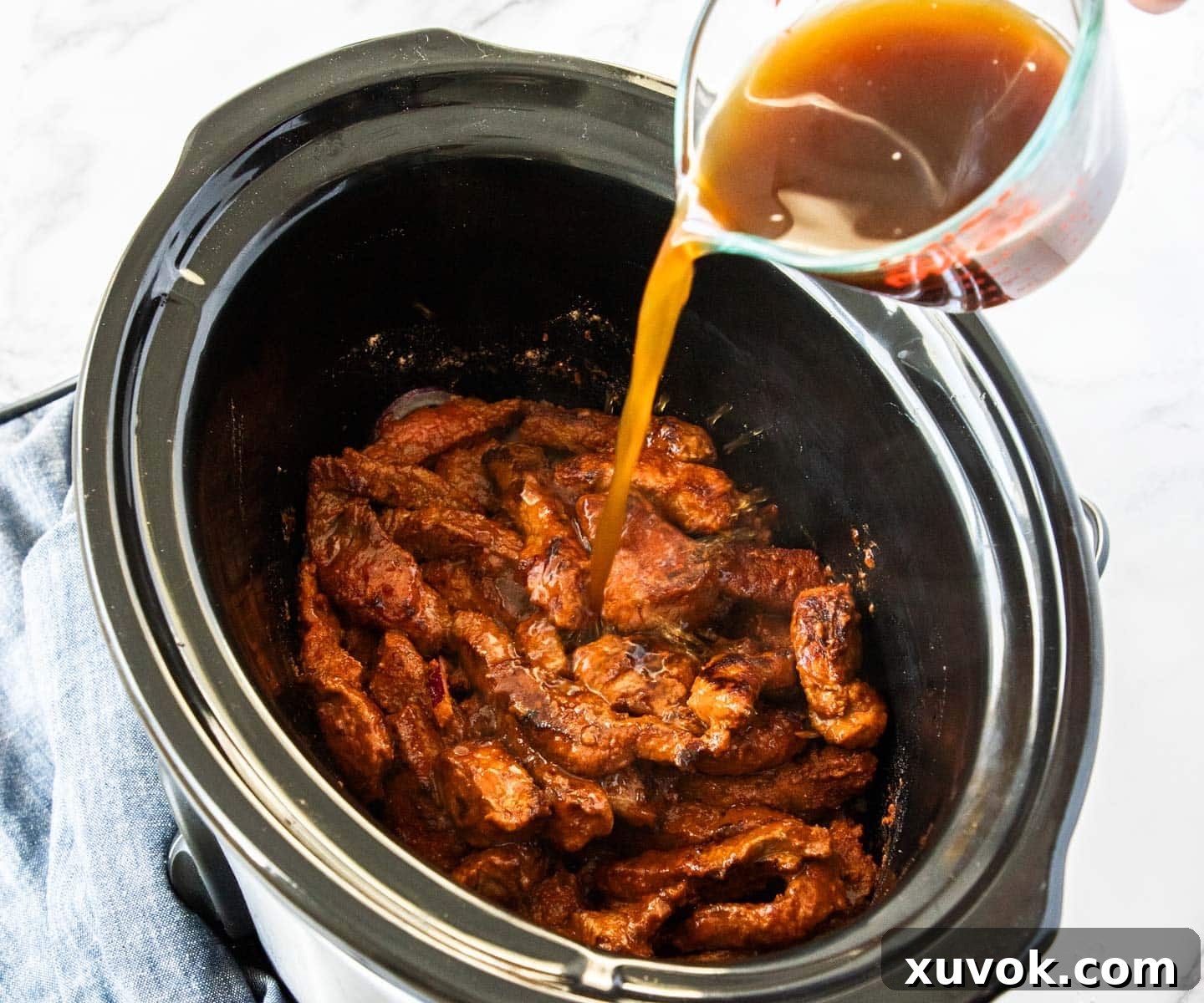Pouring beef stock over the seasoned beef and vegetables in a slow cooker bowl.