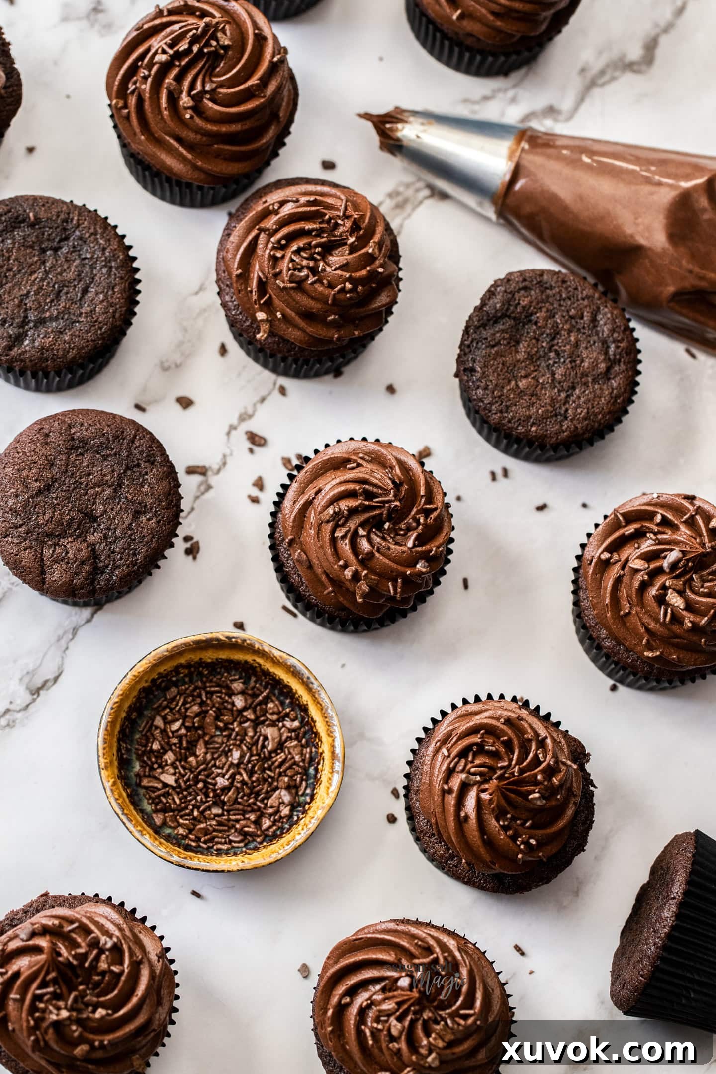 A top-down view of a dozen beautifully frosted chocolate fudge cupcakes, accompanied by a bowl of colorful sprinkles and a piping bag.