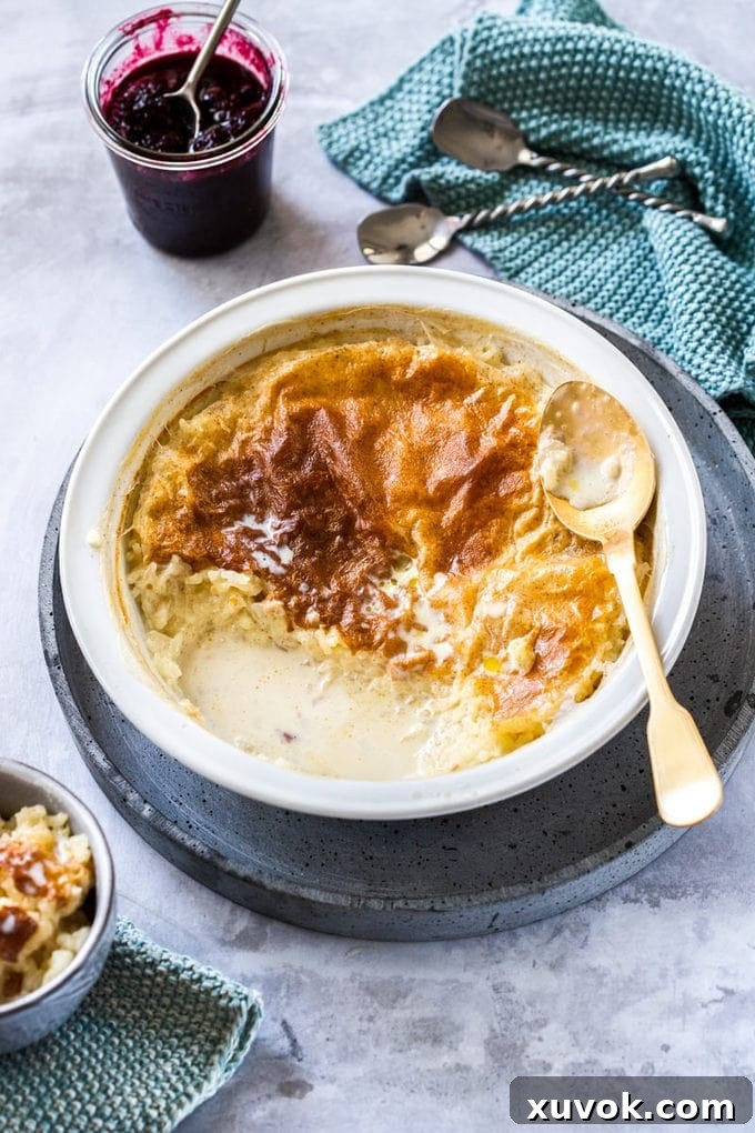 Birdseye view of a pie dish filled with rice pudding. A gold spoon on the side and a pot of blackberry compote behind it.