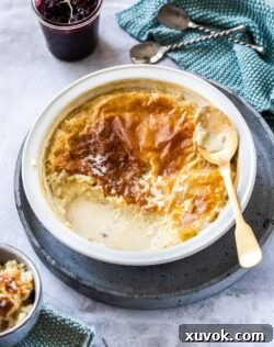 Birdseye view of a pie dish filled with rice pudding. A gold spoon on the side and a pot of blackberry compote behind it.