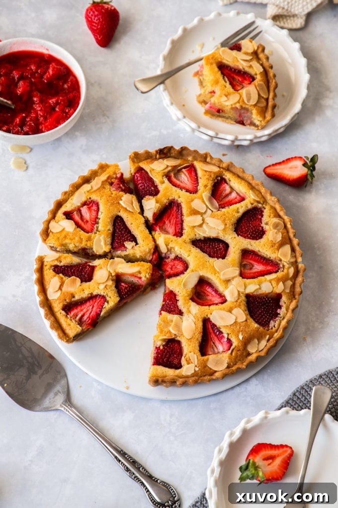 Birdseye view of a strawberry tart on a white plate. A cake slice sits in front and dessert plates are set around it.
