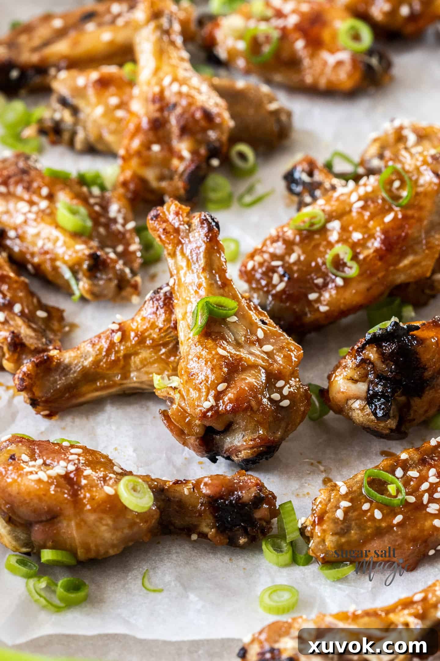 A batch of shiny, golden-brown honey soy coated chicken wings on a sheet of baking paper, garnished with sesame seeds.