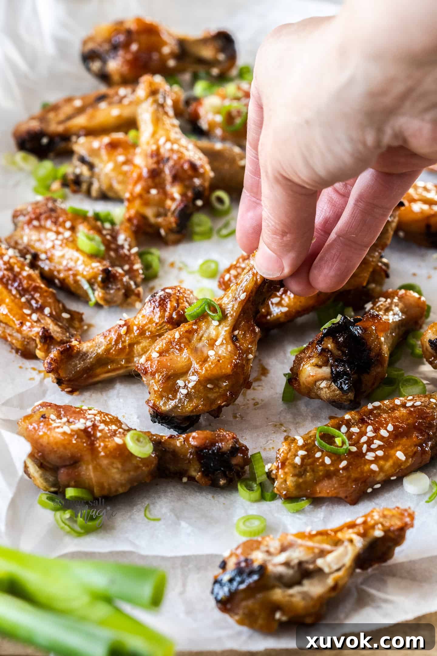A hand reaching for a perfectly cooked, glossy honey soy chicken wing on a baking sheet, highlighting its sticky texture.