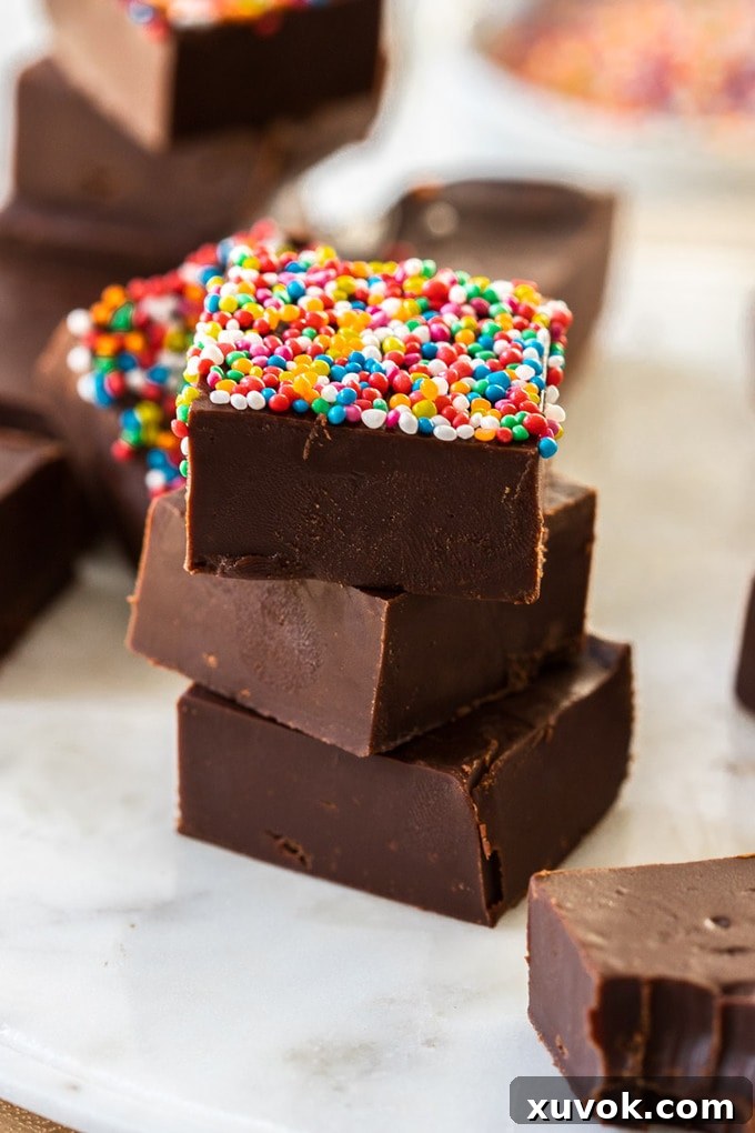 A charming stack of dark chocolate fudge squares on a white marble platter, with the top piece beautifully decorated with sprinkles, highlighting its delicious texture.