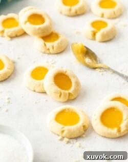 A batch of golden lemon curd cookies on a sheet of baking paper, with a small spoon of vibrant yellow lemon curd placed beside them, highlighting the delicious filling and buttery texture.