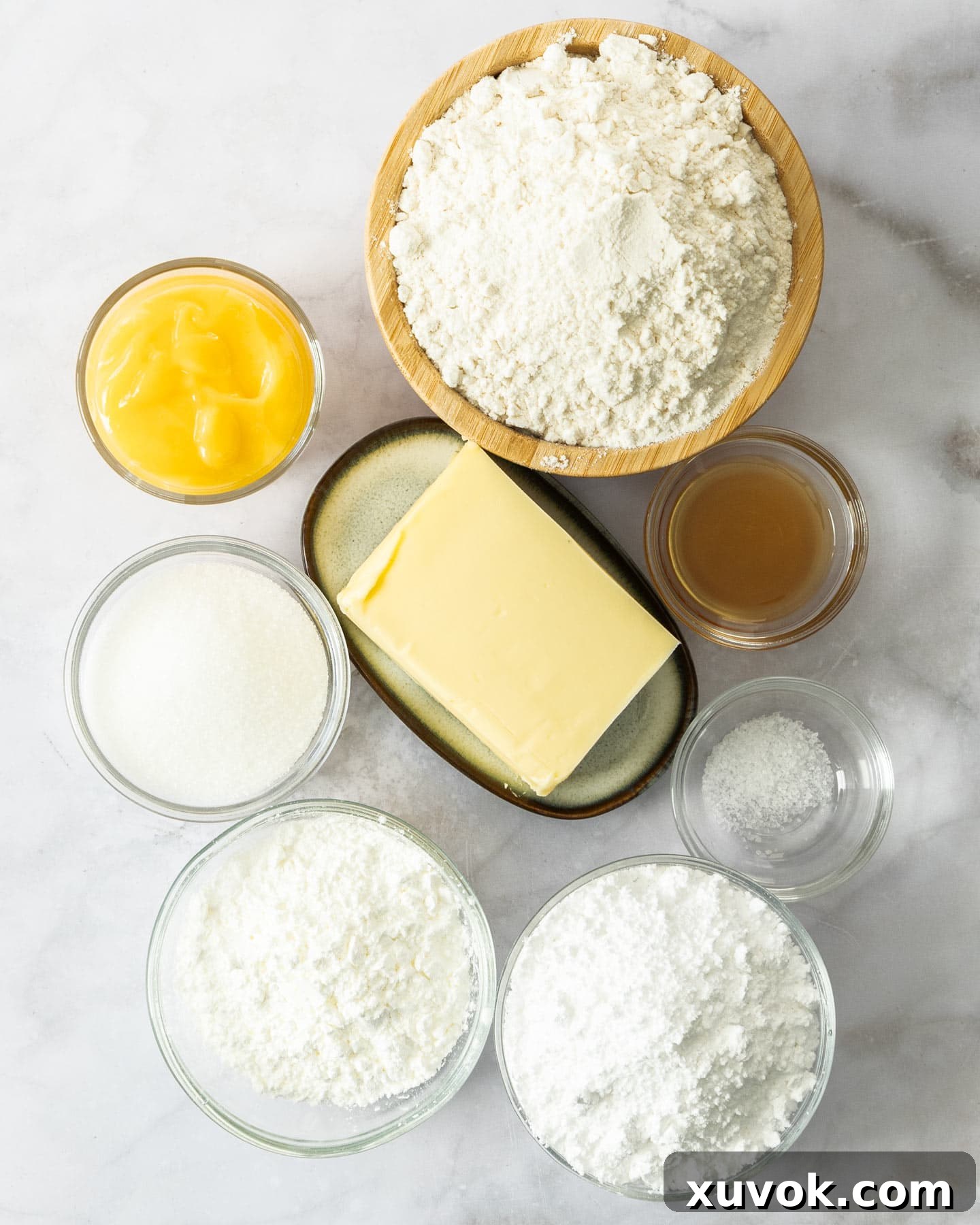 A selection of raw ingredients for lemon curd cookies, including blocks of butter, powdered sugar, flour, and a bowl of bright golden lemon curd, neatly arranged on a pristine white surface.