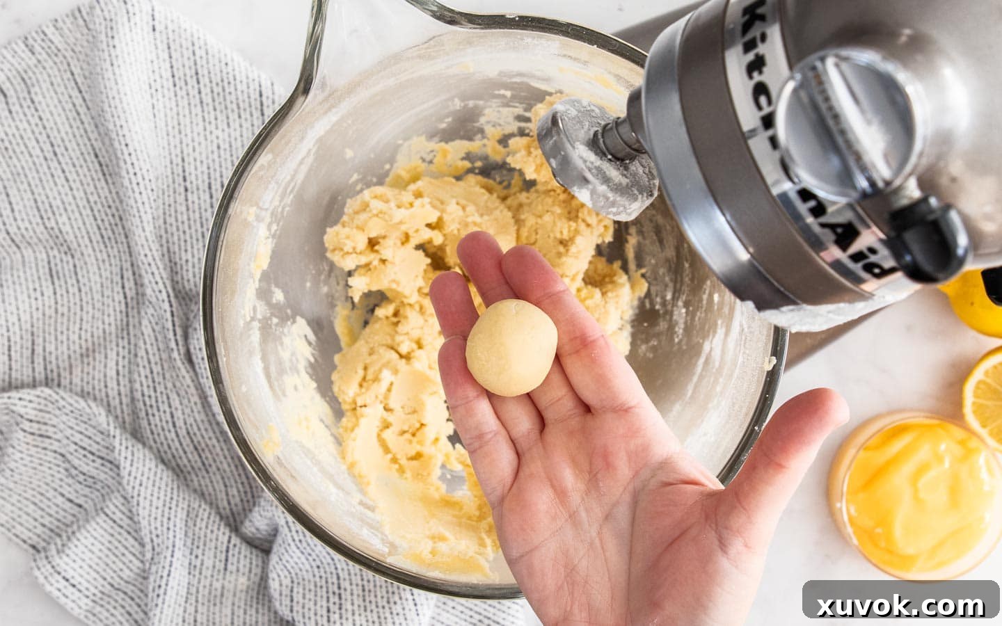 A baker's hand gently shaping a small, round ball of lemon curd cookie dough, illustrating the careful crafting involved in preparing each individual treat.