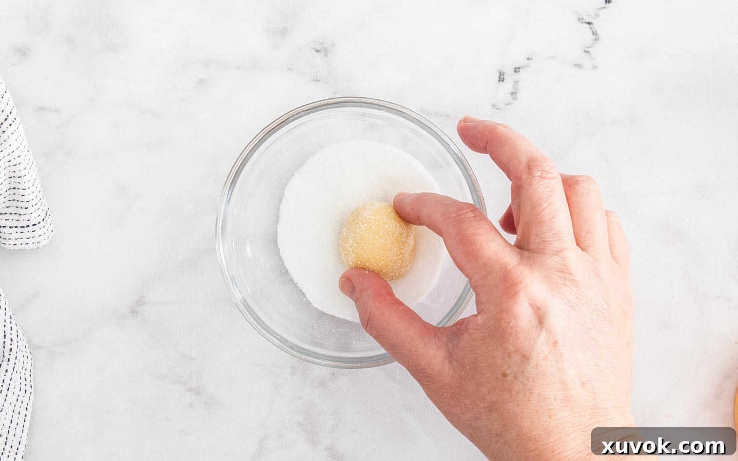 A close-up shot of a cookie dough ball being rolled in granulated sugar, showing an even coating that will add a delightful crunch and sparkling finish to the baked lemon curd cookies.